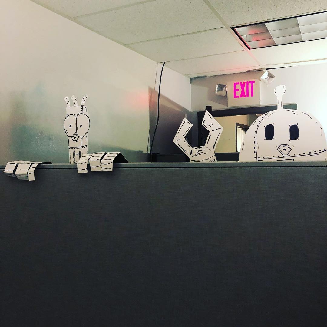 Illustrated robots cut out of carboard peeking over a cubicle.