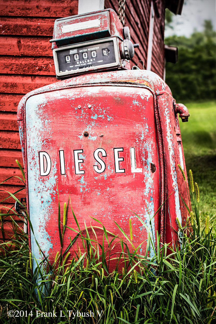 An old, faded gas pump in front of a barn. Overgrown grass surrounds it. The word "Diesel" is painted on the side.