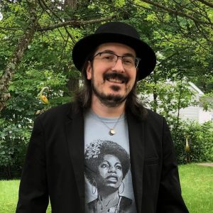 Frank Tybush, a white non-binary person. They have dark brown hair and beard, both streaked with gray. They have a black flat top, wide brimmed hat on, a black suit jacket, and a blue t-shirt with an illustration of Nina Simone.