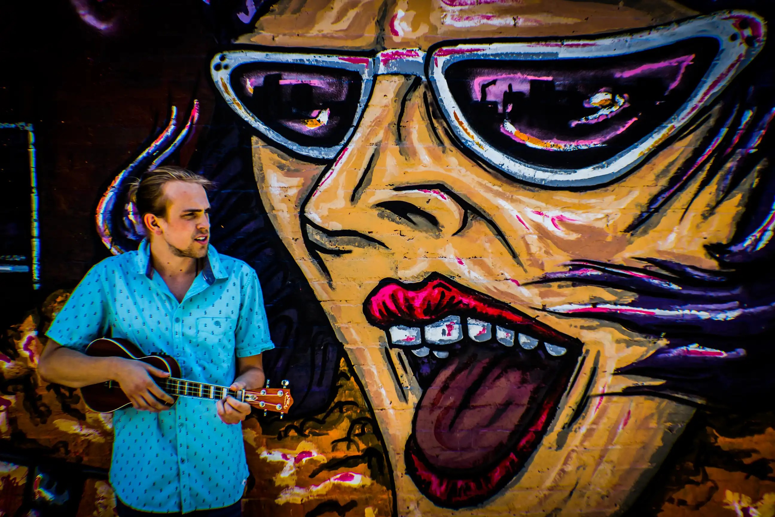 A young white man with blond hair and a scruffy beard stands in front of a mural of a woman with sunglasses and her mouth open.