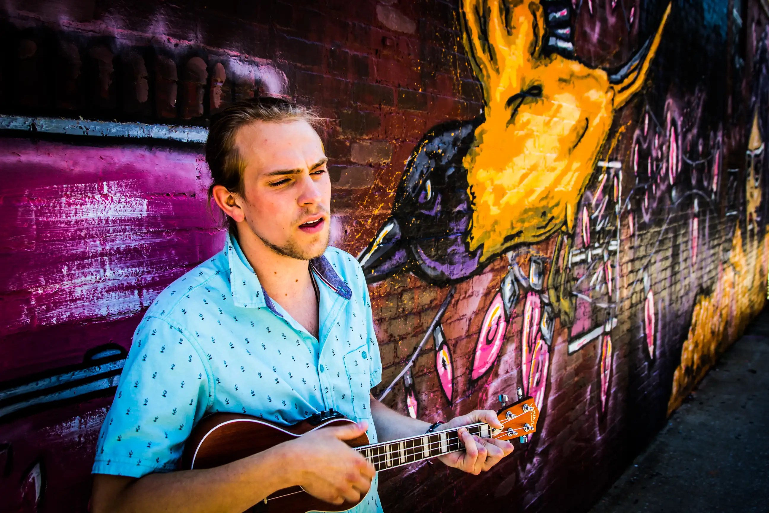 A young white man with blond hair and a scruffy beard is standing against a brick wall. He's holding a ukulele and there's a distorted mural on the wall.