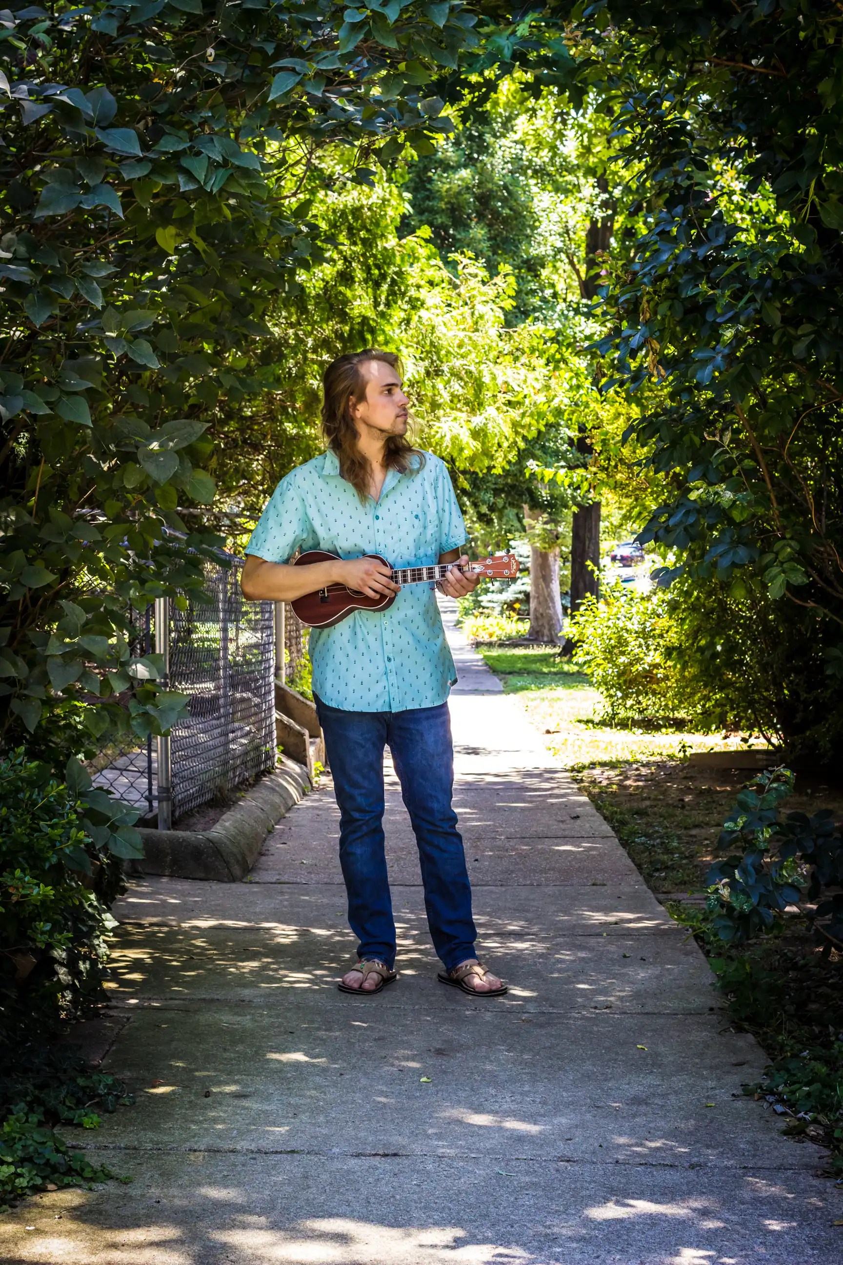 A young white man with blond hair and a scruffy beard holding a ukulele while standing under a leaf covered trellis over a sidewalk