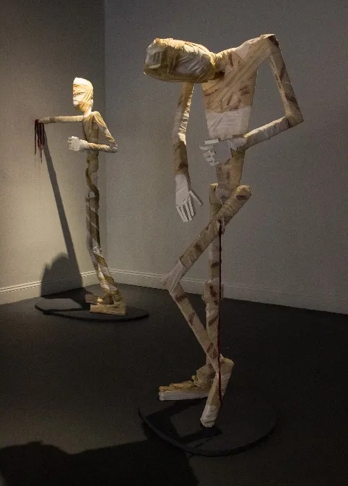 Two wooden sculptures in a gallery. They are covered in stained fabric.