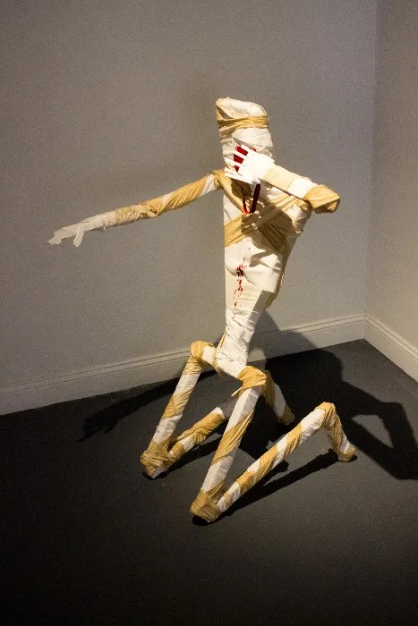 A wooden sculpture posed on its knees. It's wiping it's bloody mouth while flushing an imaginary toilet.