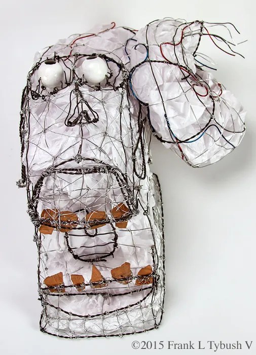 A wire sculpture of a head being wrenched back and into a coil. The scultpure is stuffed with white tissues paper and has clay teeth and painted wooden eyes.
