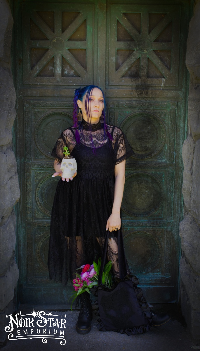 A goth woman standing in front of a Mausoleum. She's holding a skull with a flower coming out of it.