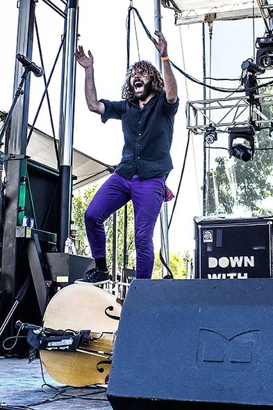 A musician standing on an upright bass, in the middle of clapping his hands. He has long hair and a beard. He's wearing a black shirt and purple pants.