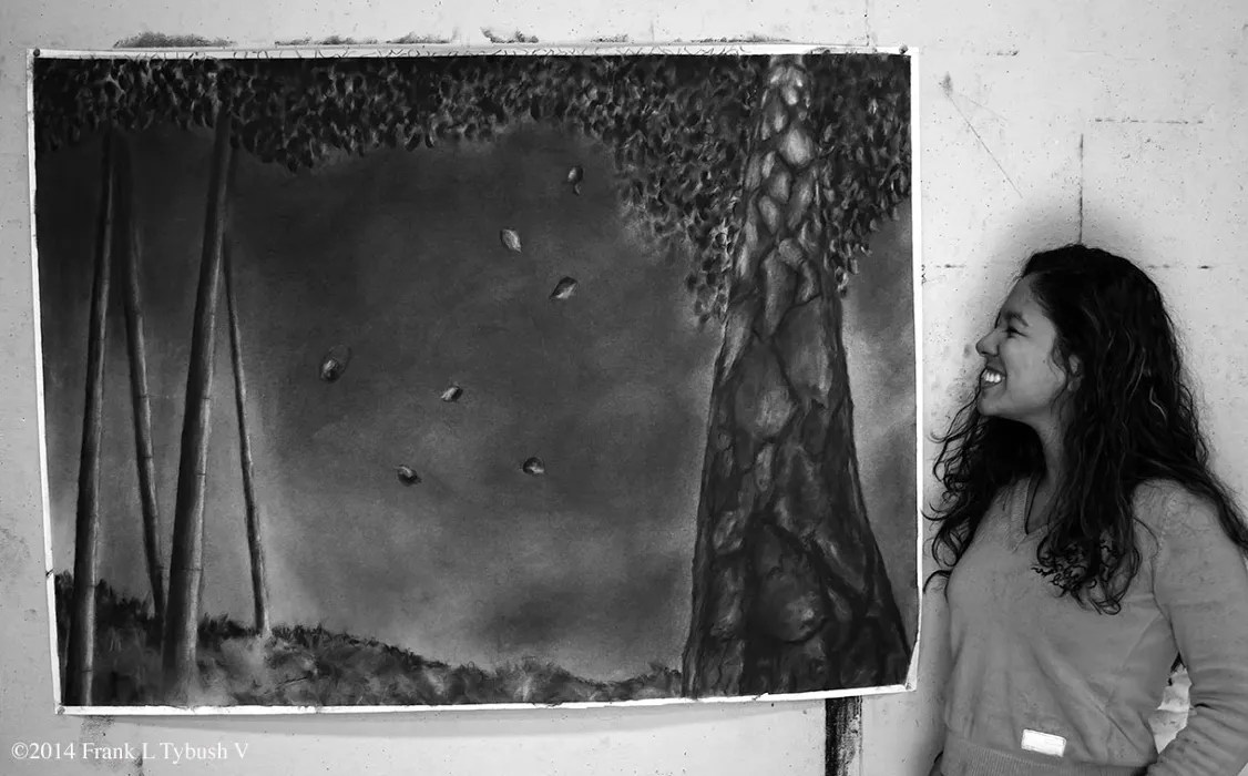 A black and white photo of a female artist smiling proudly at their charcoal drawing of a forest.