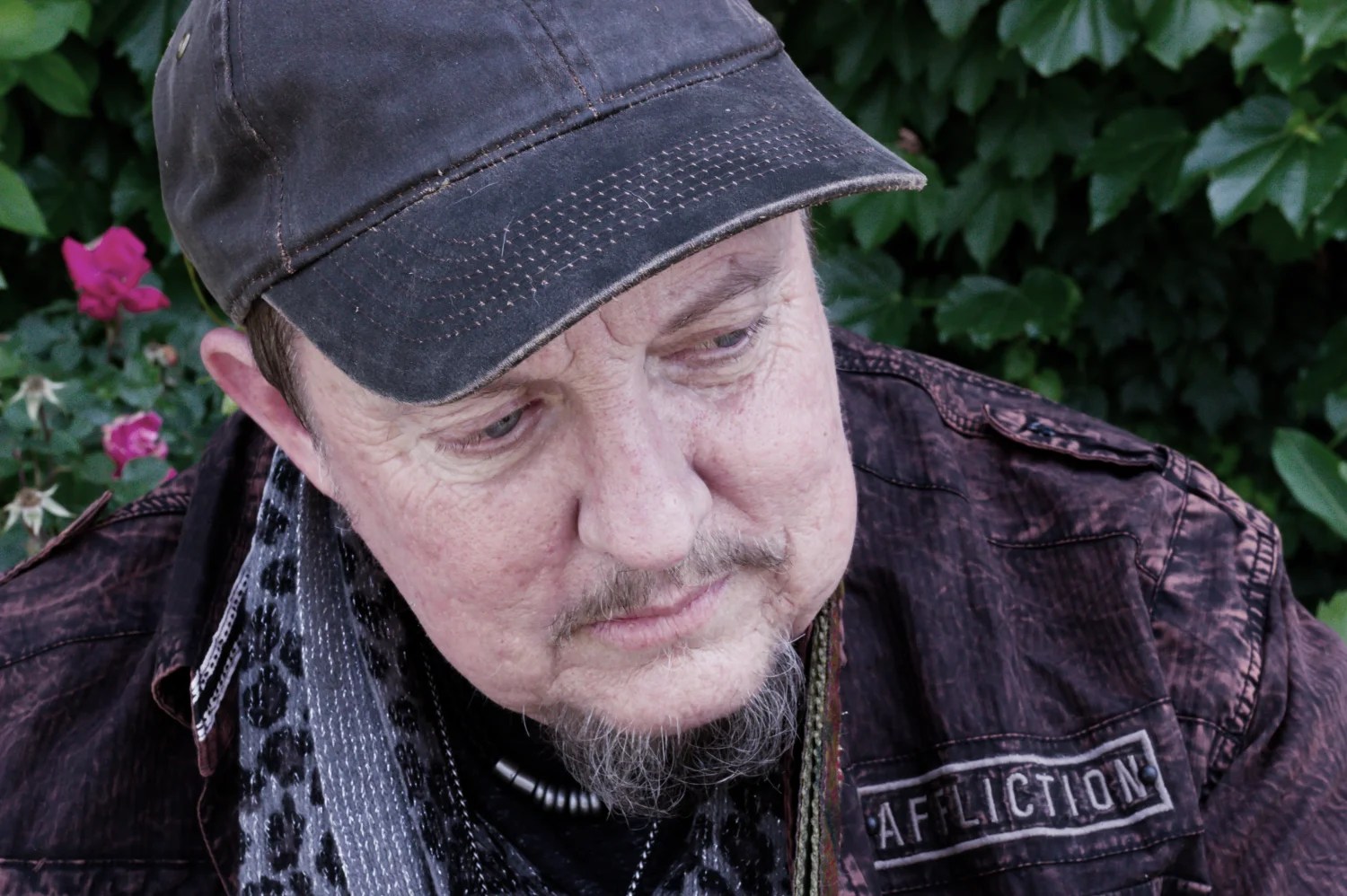 A close up on Singer/Songwriter Trace Ellis. He's wearing a black, worn baseball cap, a dark, rugged jeans jacket, a black and white leapord-print, thin scarf, and a gray t-shirt. He has a medallion around his neck.