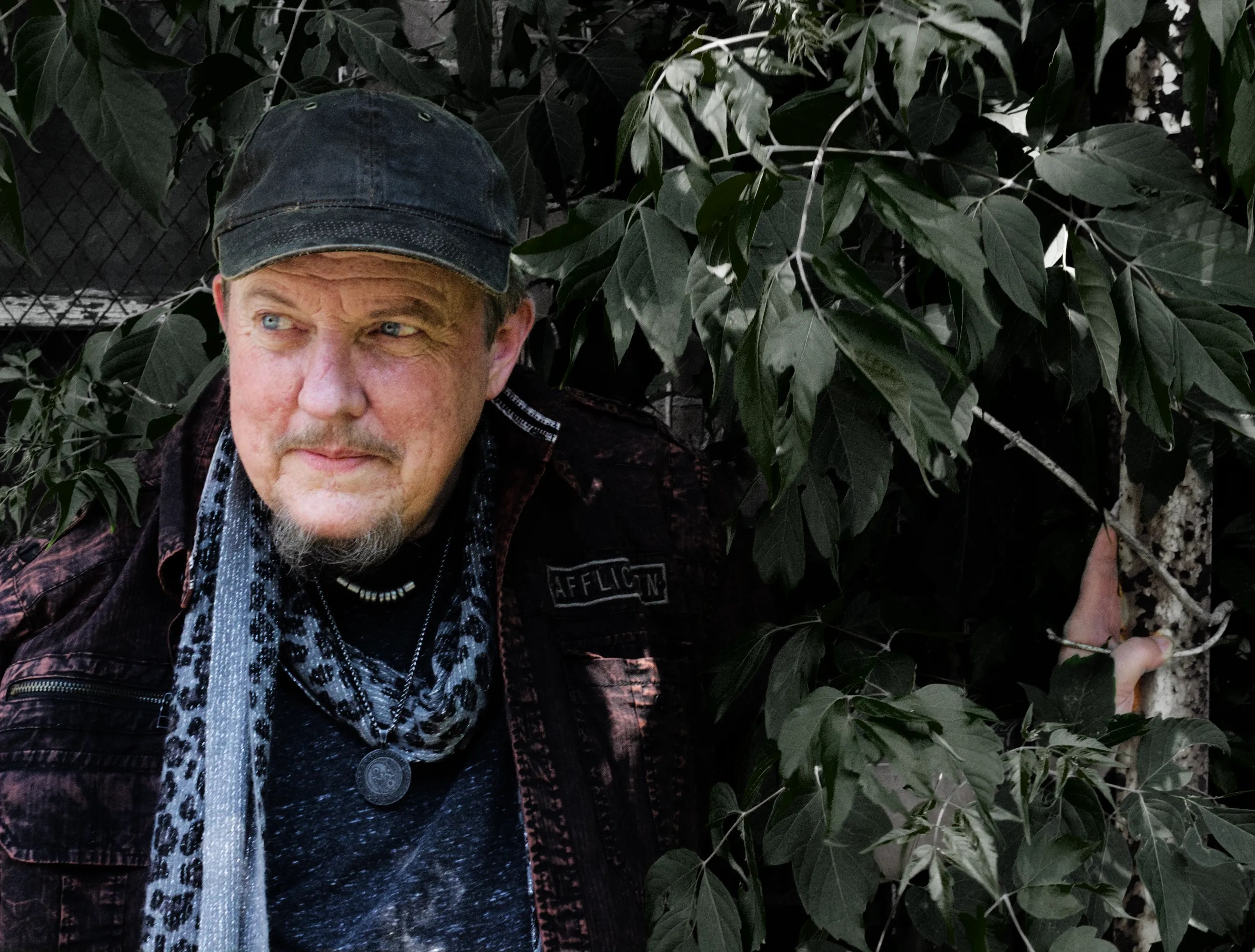 Singer/Songwriter Trace Ellis resting on an empty stage, holding his guitar. He's wearing a black, worn baseball cap, a dark, rugged jeans jacket, a black and white leapord-print, thin scarf, and a gray t-shirt. He has a medallion around his neck. He has dark pants on and brown workboots. 